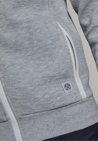 Sudadera gris con cremallera, hecha de una tela suave, que presenta detalles en blanco en la cremallera, bolsillos laterales y una etiqueta de la marca en la parte inferior izquierda.