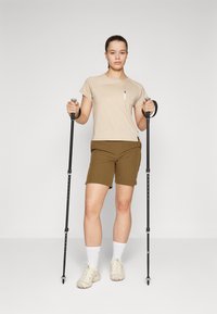 Beige kortermet skjorte med frontlomme, brune shorts og lyse sko; holder sorte turtøfler; ensfarget bakgrunn.