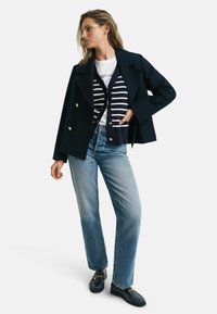 Marineblazer mit doppelter Knopfleiste über einem marine- und weiß gestreiften Cardigan, kombiniert mit hellblauen Jeans und dunklen Loafers mit Kettendetail.