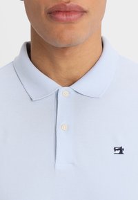 Vaaleansininen polo-paita, jossa on tekstuuri pintakäsittely. Paita on varustettu klassisella kauluksella, kahdella beigellä napilla ja pienellä mustalla ompelukonelogolla rinnassa.