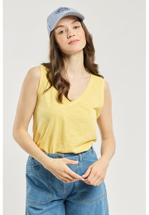 Jeune femme aux longs cheveux bruns portant une casquette rayée bleue, un top jaune sans manches et un jean bleu taille haute sur un fond uni.