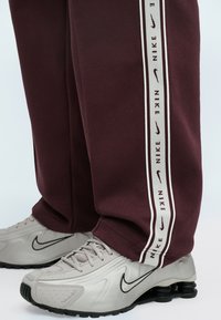 Bordo treniņbikses ar baltām sānu svītrām, uz kurām ir "Nike" zīmols. Pāris ar pelēkiem sporta apaviem, kas ir ar teksturētu dizainu un izliekumu.
