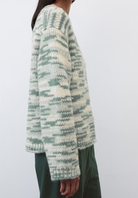 Donna che indossa un maglione a maglia spessa con motivi astratti verdi e bianchi, abbinato a pantaloni verde scuro, in piedi contro un muro semplice.