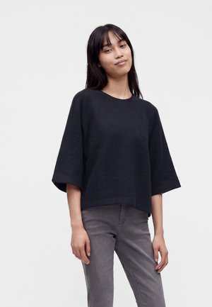 Jeune femme aux longs cheveux foncés porte un pull noir ample à manches jusqu'aux coudes et un jean gris sur un fond clair uni.