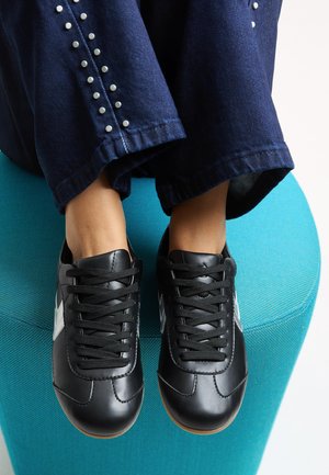 Sneaker in pelle nera con lacci neri e una striscia bianca. Posizionati su un sedile turchese, abbinati a pantaloni di jeans scuri con borchie argentate.
