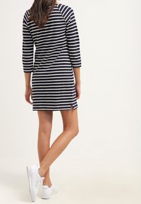 Femme portant une robe mi-longue à rayures noires et blanches avec des manches trois-quarts, debout les jambes croisées, portant des baskets blanches.