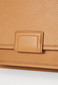 La borsa in pelle marrone chiaro in primo piano presenta una superficie testurizzata con una chiusura metallica rettangolare e dettagli cuciti, evidenziando una finitura elegante.