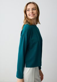Pullover lungo a maniche lunghe in teal, realizzato in materiale morbido, con piccole strass sparse come dettagli, dal taglio rilassato e con fessure laterali.