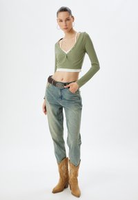 Grünes geripptes Crop-Top mit langen Ärmeln, hochgeschnittene hellblaue Jeans, brauer Gürtel und beige Cowboy-Stiefel mit Stichdetails.