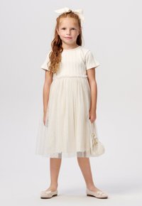 Romantische jurk van crème met een ribbelstructuur aan de bovenkant en een tule rok, verfraaid met sparkles. Gepaard met zachte ballerina's en een kleine handtas.