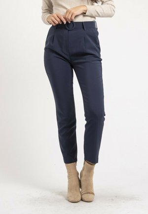 Femme attachant une ceinture sur un pantalon bleu marine fuselé, portant des bottines en daim beige et un haut à manches longues beige clair sur un fond uni.