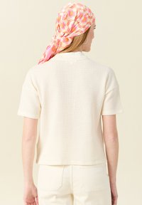 Kortärmad, krämfärgad stickad topp med en strukturerad väv, tillsammans med en rosa blommig halsduk. Toppens passform är avslappnad och den har en något kortad längd.