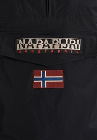 Czarna kurtka z tkaniny, na której znajduje się wyraźna, tkana etykieta "Napapijri geographic" w kolorach beżowym i pomarańczowym, a poniżej naszywka z czerwonym norweskim flagą.