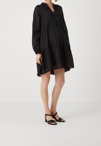 Vestido negro de ojales con mangas largas, escote en pico y falda con volantes. Combinado con sandalias de tacón negro con tiras sobre un fondo neutro.