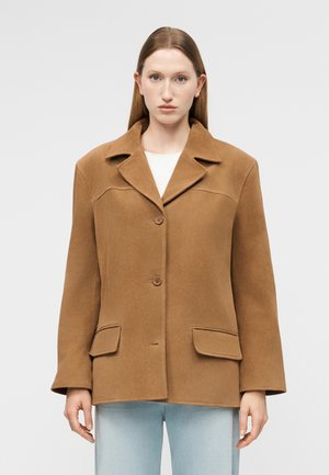 Γυναίκα με ίσια μαλλιά που φοράει ένα κουμπωτό καμηλό καφέ παλτό από μαλλί με φαρδιούς γιακάδες και τσέπες με καπάκι, συνδυασμένο με ανοιχτόχρωμο μπλε τζιν.