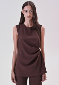 Donna con lunghi capelli scuri che indossa un top senza maniche marrone con dettagli in fibbia laterale e pantaloni marroni abbinati su uno sfondo semplice.