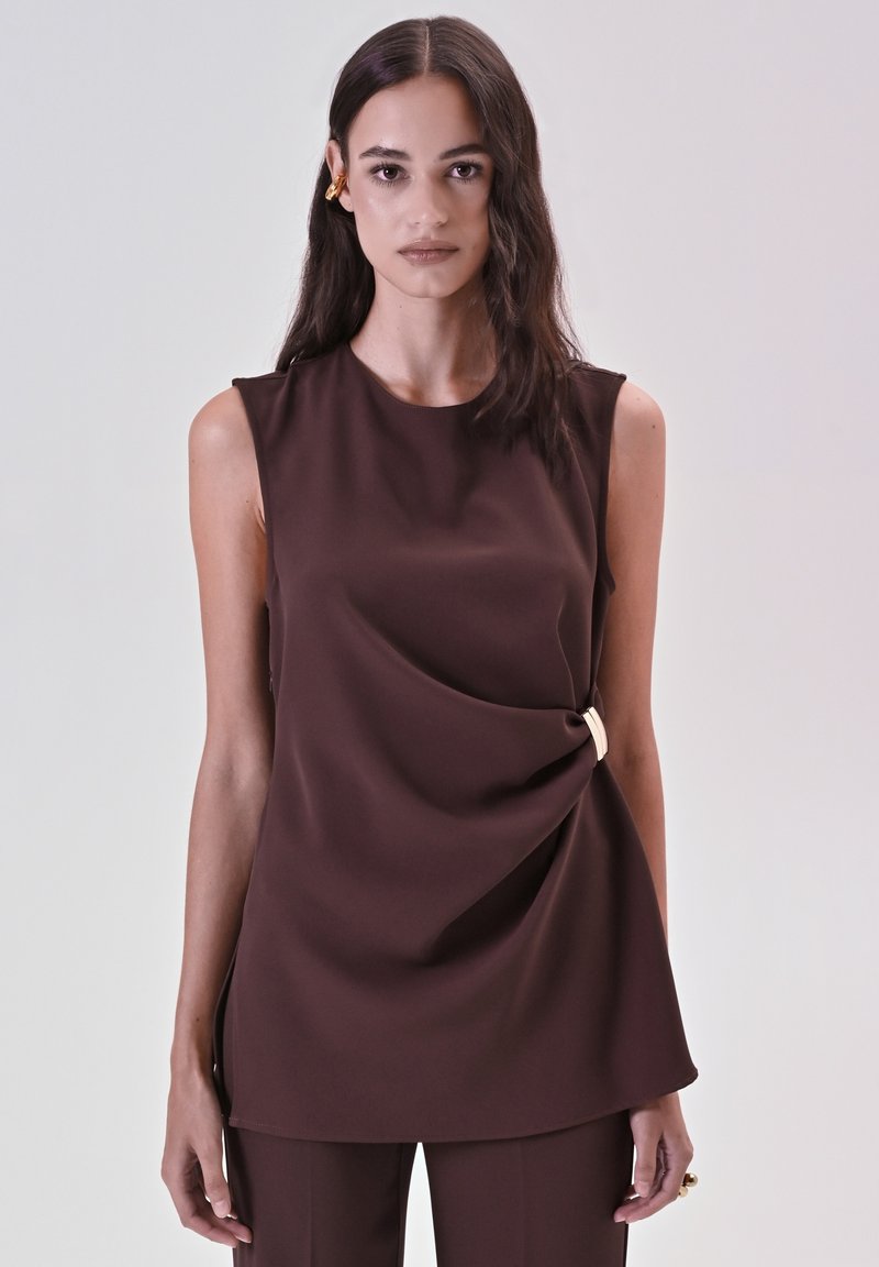 Donna con lunghi capelli scuri che indossa un top senza maniche marrone con dettagli in fibbia laterale e pantaloni marroni abbinati su uno sfondo semplice.