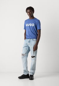 Kék pamut póló fehér "HUGO" felirattal. Világoskék, tépett szárú farmernadrág, szálkás széllel. Fekete, sima felületű utcai cipő.