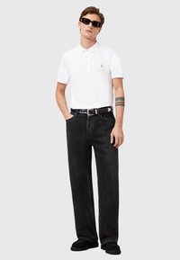 Witte polo met korte mouwen, zwarte hoge taille jeans, zwarte riem met zilveren hardware, zwarte platte schoenen, gedragen met zwarte zonnebril.