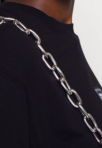 Zwarte sweat met een grove zilveren kettingband en een klein merkpatch. De stof lijkt zacht met een geribbelde halslijn.