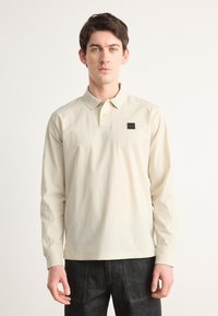 Langermet polo-skjorte i lys beige bomull med krage og et lite svart merke på venstre side. Har en avslappet passform.