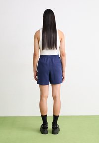 Námornícké modré kraťasy s elastickým pasem, spárované s béžovým tílkem. Osoba má dlouhé, rovné vlasy a nosí černé ponožky a boty.
