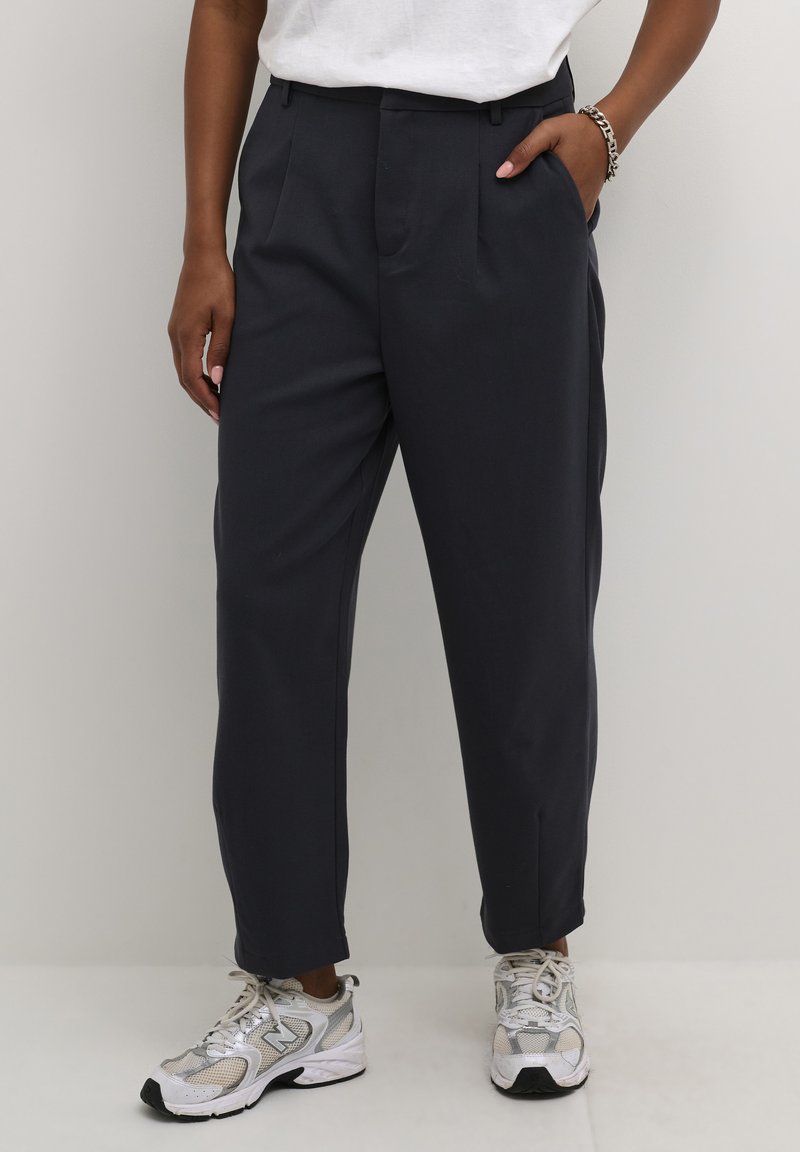 Pantalon taillé gris foncé avec plis, présentant une coupe droite. Associé à une chemise blanche et des baskets grises. Texture de tissu lisse.