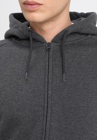 Sudadera gris con cremallera frontal, capucha con cordones y textura suave. Presenta un diseño informal adecuado para usar en capas.