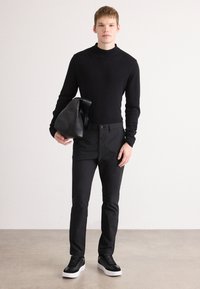Suéter de cuello alto negro, pantalones ajustados negros y zapatillas negras con suelas blancas. Sostenido un bolso negro texturizado. Fondo neutro.
