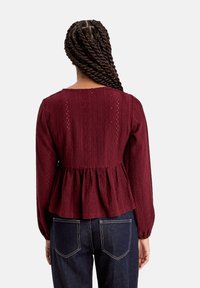Haut peplum bourgogne à manches longues, en tissu texturé avec des motifs en dentelle élaborés, col rond et détail de taille froncée.