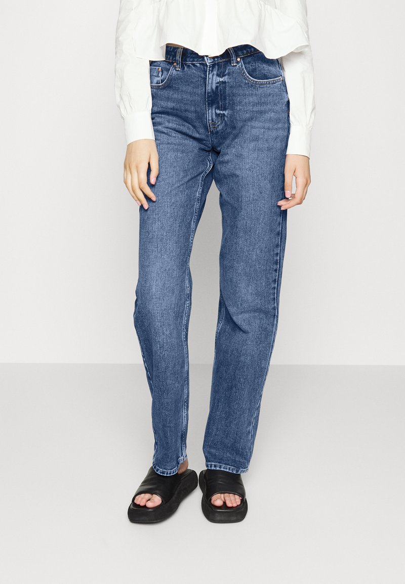 Jeans de ganga azuis com perna reta, design de cinco bolsos e cintura média. Usados com uma camisa branca cropped e sandálias pretas.