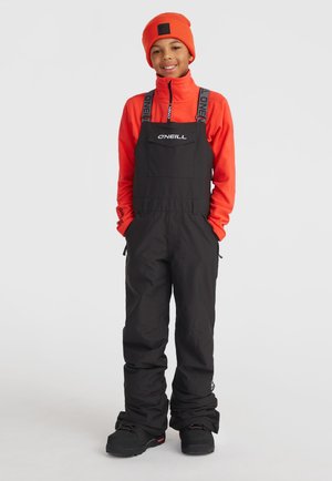 Salopette de neige noire avec des bretelles réglables, comportant une poche avant. Portée par-dessus un polaire orange vif, complétée par un bonnet assorti.