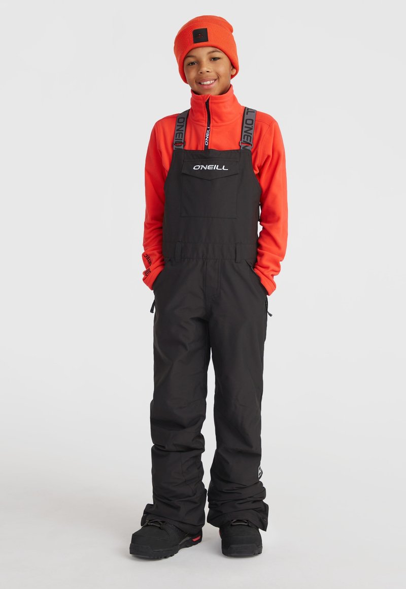 Čierne snehové overall s nastaviteľnými popruhmi, so prednou kapsou. Nosené cez jasne oranžové fleece, doplnené zodpovedajúcou čiapkou.