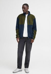 Fleece dzseki magas gallérral, zöld és tengerészkék színblokkokkal, elülső cipzáros zsebbel, fehér inggel és sötét farmernadrággal viselve.