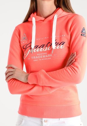 Osoba ubrana w koralową bluzę z kapturem z białymi ściągaczami i logo żeglarskiej marki "Gaastra", z założonymi skrzyżowanymi ramionami, w połączeniu z białymi spodniami.