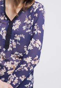 Chemisier en dentelle florale bleu marine avec des fleurs beige clair, manches longues et un décolleté en V noir. Tissu léger avec une texture douce.