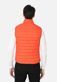 Gilet imbottito arancione con cuciture verticali, colletto alto e una texture leggera, indossato sopra una camicia a maniche lunghe bianca. Pantaloni neri visibili.