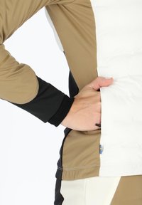 Hybridjacke mit beigen, schwarzen und weißen Paneelen. Glatte Textur mit kontrastierenden Farben und einer dezenten Tasche an der Seite.