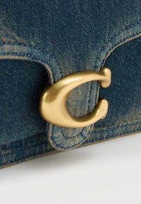 Κοντινή λήψη χρυσού μεταλλικού κλεισίματος σε σχήμα "C" προσαρτημένου σε μπλε τζιν ύφασμα με ορατή ραφή.