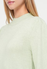 Gaiši zaļš džemperis ar augstu apkakli, ribota tekstūra un pūkaina piedurkne. Mīksta adīta auduma ar maigu šuvju detaļu gar malām.