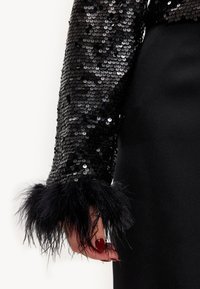 Manche noire à sequins avec manchette noire à plumes, design ajusté, texture lisse, associée à une robe noire unie, mettant en valeur les détails et le contraste.