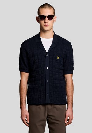 Uomo che indossa una camicia nera a maniche corte con bottoni e motivo testurizzato, con logo di un uccello giallo, maglietta bianca sotto, pantaloni marroni e occhiali da sole scuri, su sfondo grigio.