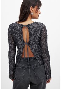 Črna majica z dolgimi rokavi, okrašena s cekini in z odprtim hrbtom, zavezana z sivo ribon. Ima oprijet kroj in teksturirano tkanino. V kombinaciji s temno jeans hlačami.