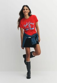 Sarkana kokvilnas T-krekls ar grafisku tekstu "Feel Good Run Club," kombinēts ar melnu svārku un melniem ceļgalu augstiem zābakiem ar baltu apmali.