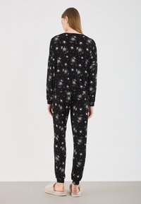 Ensemble de pyjama noir avec un motif de formes étoilées blanches. Le haut a des manches longues et le pantalon a des poignets élastiques. Accompagné de pantoufles beiges.
