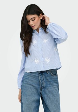 Femme ajustant ses cheveux, portant une chemise boutonnée à rayures bleu clair et blanches avec une broderie florale blanche, et un jean bleu taille haute.