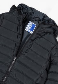 Chaqueta de plumas negra con capucha, que presenta un detalle de acolchado horizontal, un cierre frontal y una etiqueta azul en el interior. Textura suave y brillante.