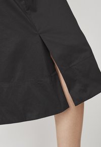 Gonna nera con uno spacco laterale, realizzata in un tessuto liscio. L'orlo è curvo e presenta un dettaglio di cucitura uniforme lungo il bordo.