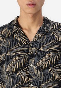 Camisa de manga corta de tela negra con un estampado de hojas de palma en beige y gris, con cuello y detalles de botones.