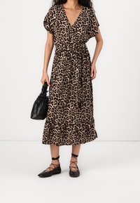 Robe mi-longue à imprimé léopard avec décolleté en V. Cette robe présente des manches courtes et une taille nouée, accompagnée de chaussures plates noires et d'un sac à main noir.